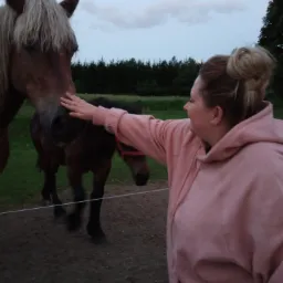 Kobieta w r&oacute;żowej bluzie głaszcze konia na pastwisku w pochmurny dzień, w tle widoczny źrebak.