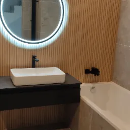 Nowoczesna łazienka z drewnianą ścianą, okrągłym lustrem LED, białą umywalką na czarnej szafce i wanną obłożoną płytkami w odcieniach beżu.