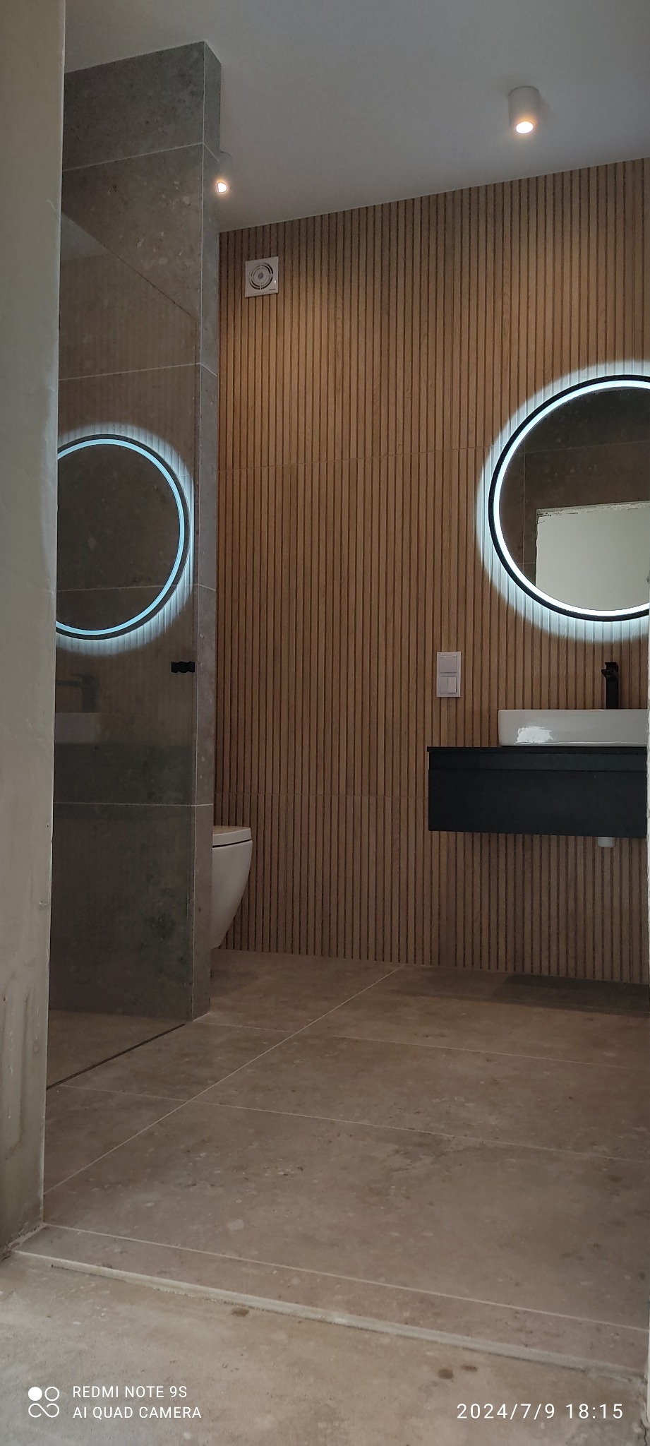 Elegancka łazienka po remoncie: drewniana boazeria, okrągłe lustra LED, podwieszana szafka z umywalką i beżowe płytki podłogowe. Minimalistyczny design.