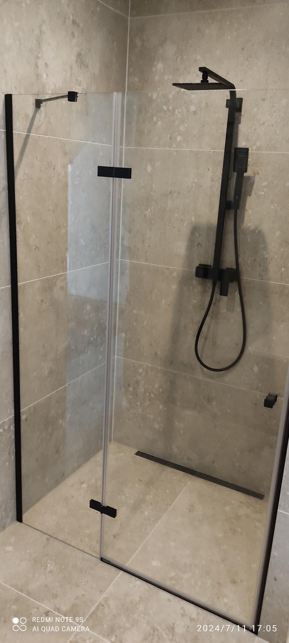 Nowoczesna kabina prysznicowa z czarnymi elementami, szklaną ścianą i deszczownicą, na tle beżowych płytek w łazience. Minimalistyczny design.