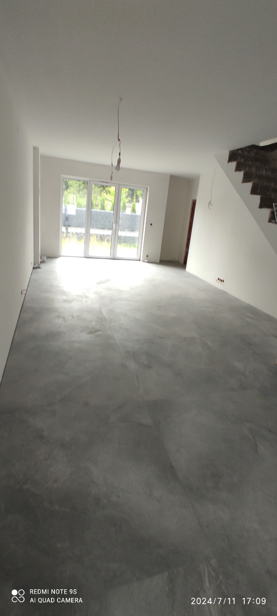 Surowa posadzka w jasnym, przestronnym wnętrzu z dużym oknem balkonowym i schodami w tle. Widoczne niedokończone elementy instalacji elektrycznej.