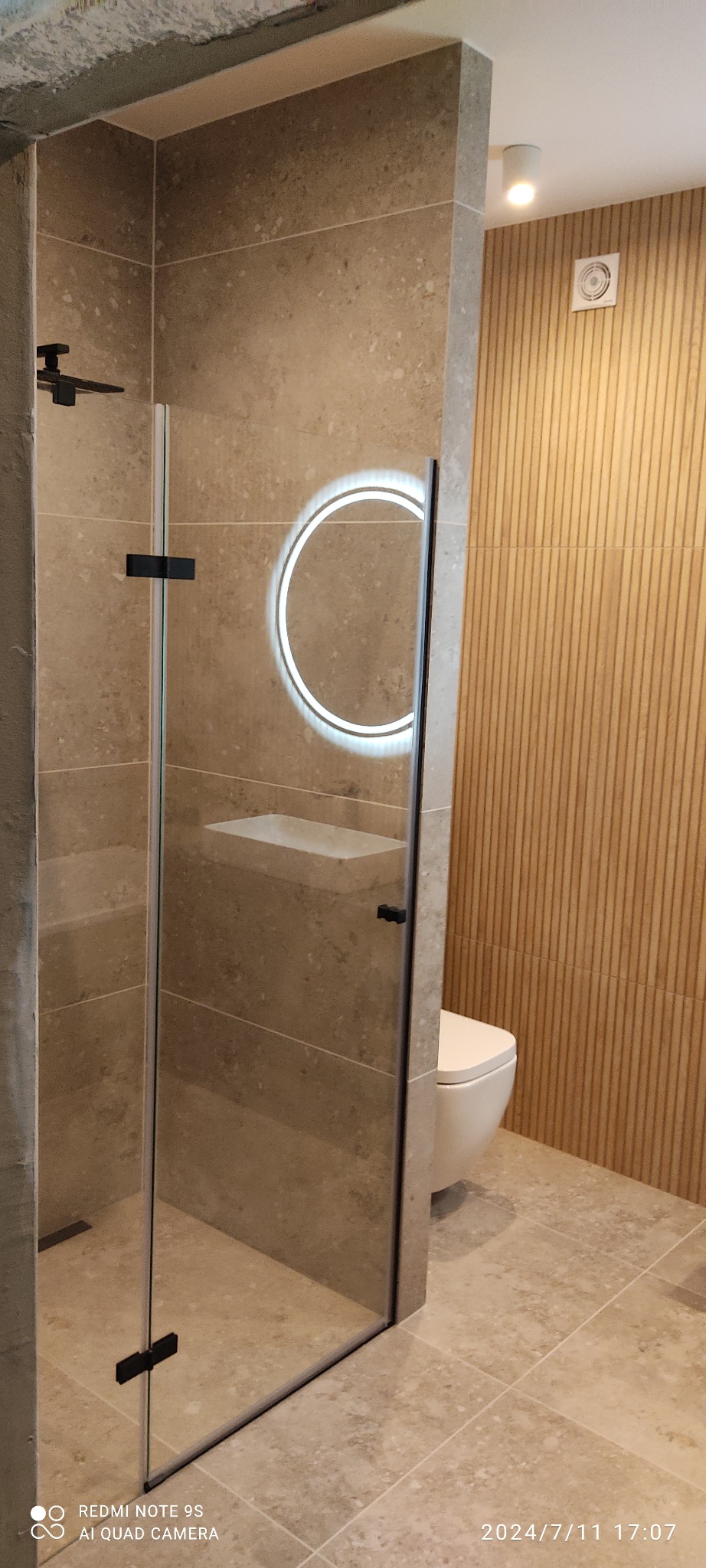 Nowoczesna łazienka z kabiną prysznicową typu walk-in, podświetlanym lustrem i beżowymi płytkami imitującymi kamień. Ściana z dekoracyjnymi panelami pionowymi.