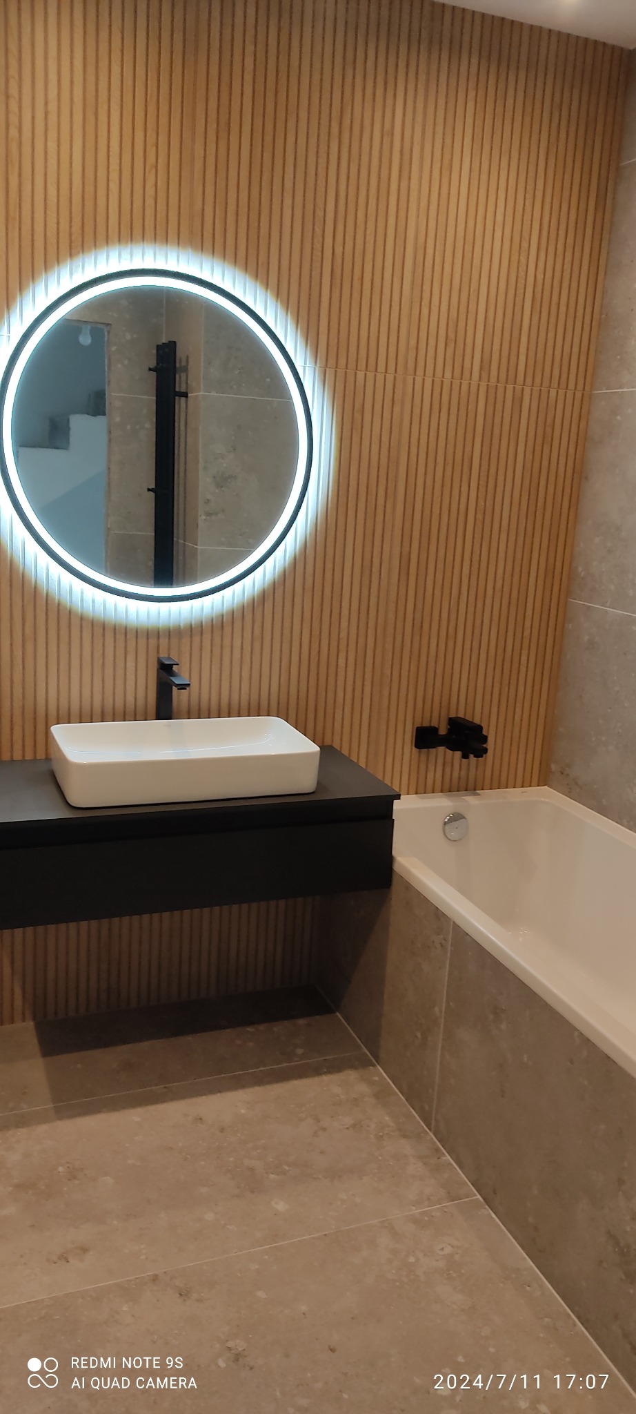 Nowoczesna łazienka z drewnianą ścianą, okrągłym lustrem LED, białą umywalką na czarnej szafce i wanną obłożoną płytkami w odcieniach beżu.
