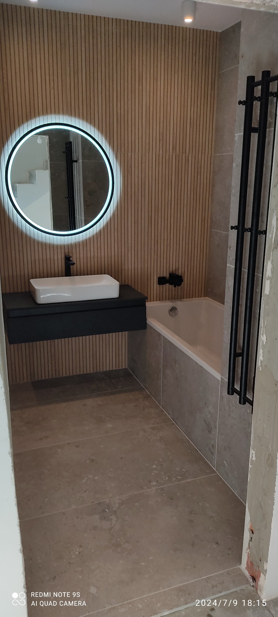 Wykończona łazienka z drewnianą ścianą, okrągłym lustrem z podświetleniem LED, czarną szafką z białą umywalką i grzejnikiem drabinkowym w kolorze czarnym.