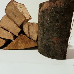 KMMOBILI Krystian Miętka - Stos pociętego drewna opałowego z widocznymi słojami, obok pionowy kawałek drewna z korą, na białym tle.