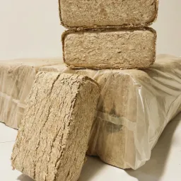 Stos ułożonych kostek brykietu drzewnego z widoczną teksturą włókien drzewnych, część zapakowana w przezroczystą folię.