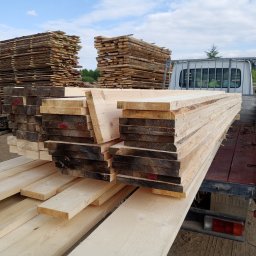 KMMOBILI Krystian Miętka - Transport drewna: deski ułożone na pace samochodu dostawczego, w tle stosy tarcicy i zalesione wzgórza pod błękitnym niebem.