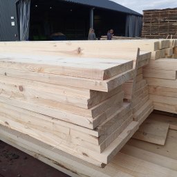 KMMOBILI Krystian Miętka - Ułożone deski z jasnego drewna na tle magazynu, widoczne osoby i stosy desek. Selekcjonowane drewno budowlane, gotowe do transportu i dalszej obróbki.