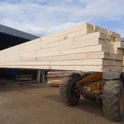 Żółty wózek widłowy transportuje stos jasnych, świeżo obrobionych desek. W tle widoczny skład drewna pod zadaszeniem i stosy desek.