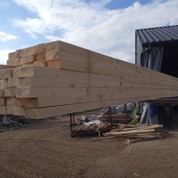 KMMOBILI Krystian Miętka - Stos belek z jasnego drewna na zewnątrz, z widocznym magazynem w tle. Niektóre belki ułożone na ziemi. Dzień słoneczny.