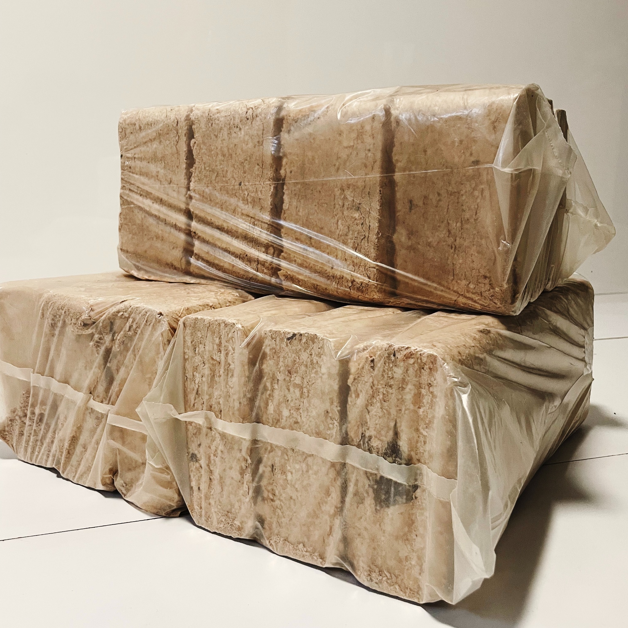 Stos trzech paczek brykietu drzewnego, owiniętych w przezroczystą folię, ułożonych na białej podłodze z widocznymi fugami.