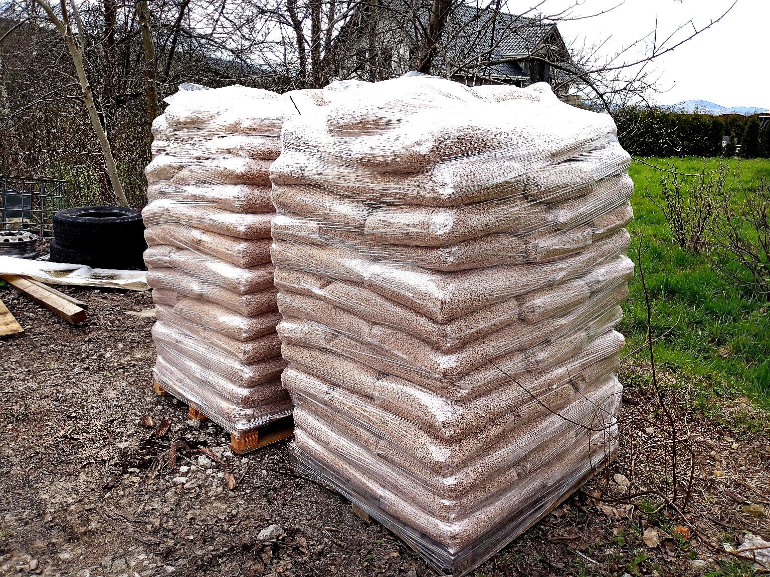 Dwa stosy worków z granulatem, zabezpieczone folią stretch, ułożone na paletach na zewnątrz, z widocznym ogumieniem w tle.