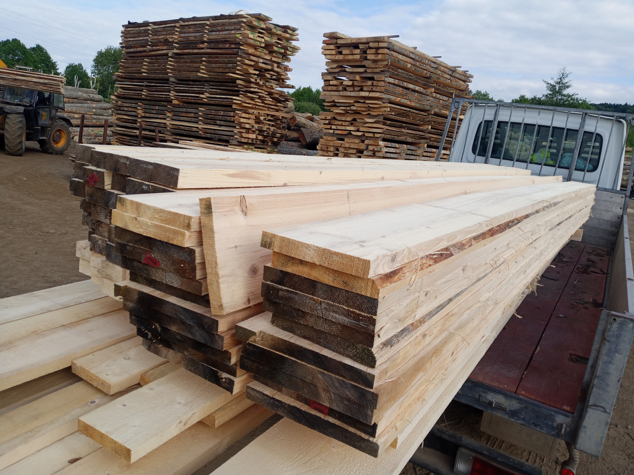 Świeżo przetarte deski ułożone w stos na pace samochodu dostawczego. W tle składowane drewno i maszyna do załadunku. Widok z tyłu pojazdu.