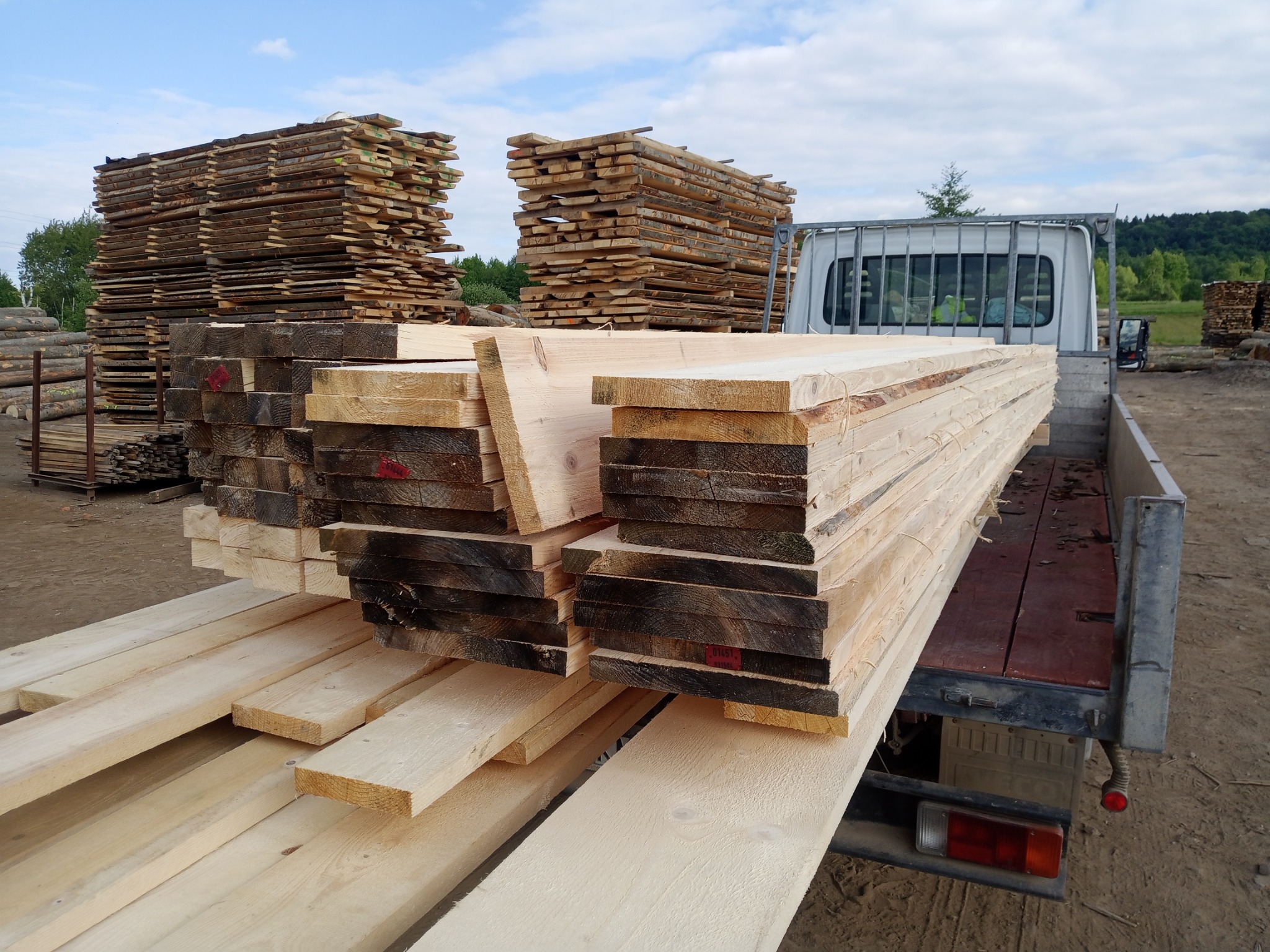 Transport drewna: deski ułożone na pace samochodu dostawczego, w tle stosy tarcicy i zalesione wzgórza pod błękitnym niebem.