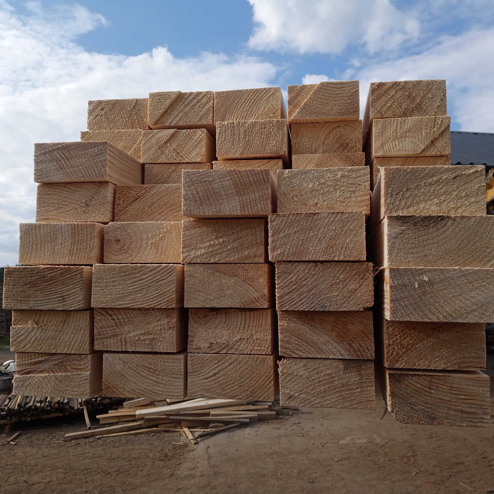 Stos równo ułożonych kantówek z jasnego drewna na zewnątrz, z widocznym niebem i chmurami w tle. Widoczne ślady cięcia i naturalny rysunek drewna.