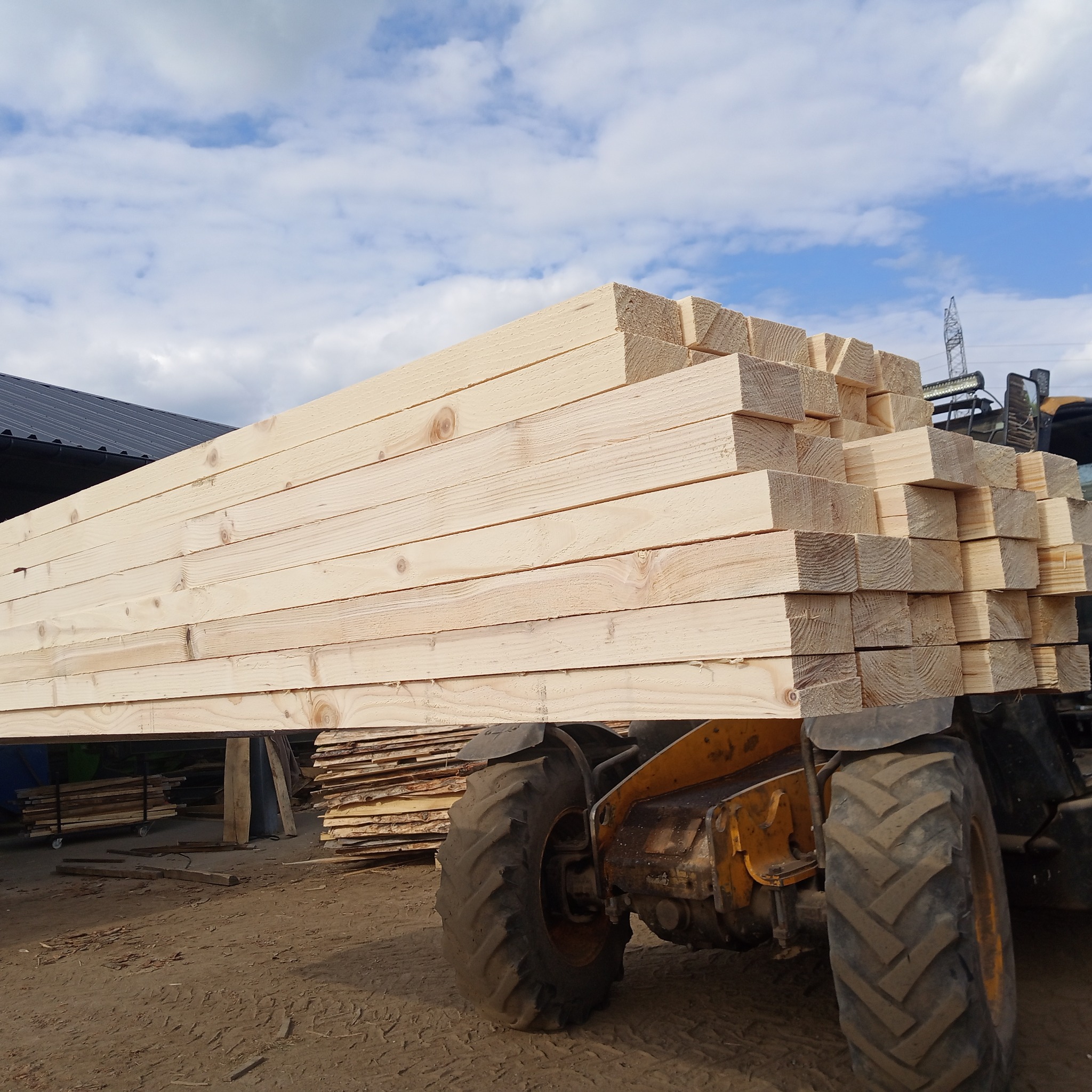 Żółty wózek widłowy przewozi stos jasnych, świeżych desek na tle błękitnego nieba z chmurami. Widoczny fragment hali i inne deski.