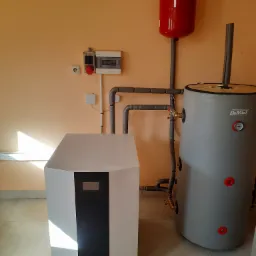 Zainstalowana gruntowa jednostka Rotorua Thermal Power z zbiornikiem akumulacyjnym i czerwonym naczyniem wzbiorczym w pomieszczeniu o beżowych ścianach, widoczne rury i osprzęt.