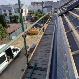 Rynna z zabezpieczeniem na dachu z rusztowaniem, widok z góry. W tle zabudowa jednorodzinna i błękitne niebo.