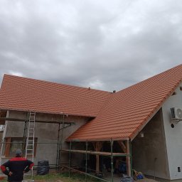 STACH-DACH CIESIELSTWO-DEKARSTWO - Dom w trakcie budowy z nowym, czerwonym dachem z dachówki. Widoczne rusztowania, drabina i pracownik z tyłu. Pochmurne niebo w tle.