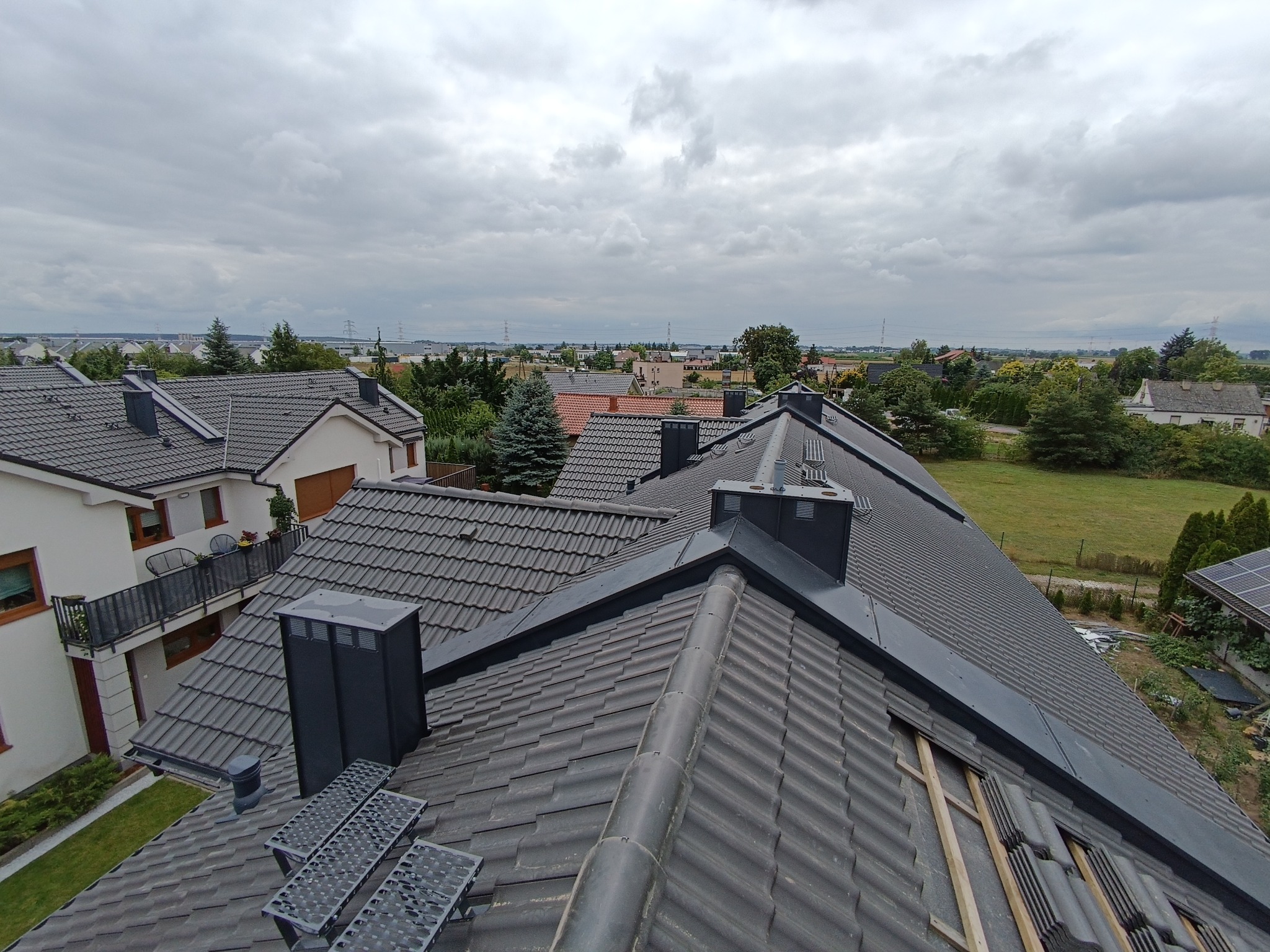 Nowoczesny dach z ciemnej dachówki ceramicznej, widok z góry na tle zabudowy jednorodzinnej i pochmurnego nieba. Fragment dachu w trakcie prac wykończeniowych.