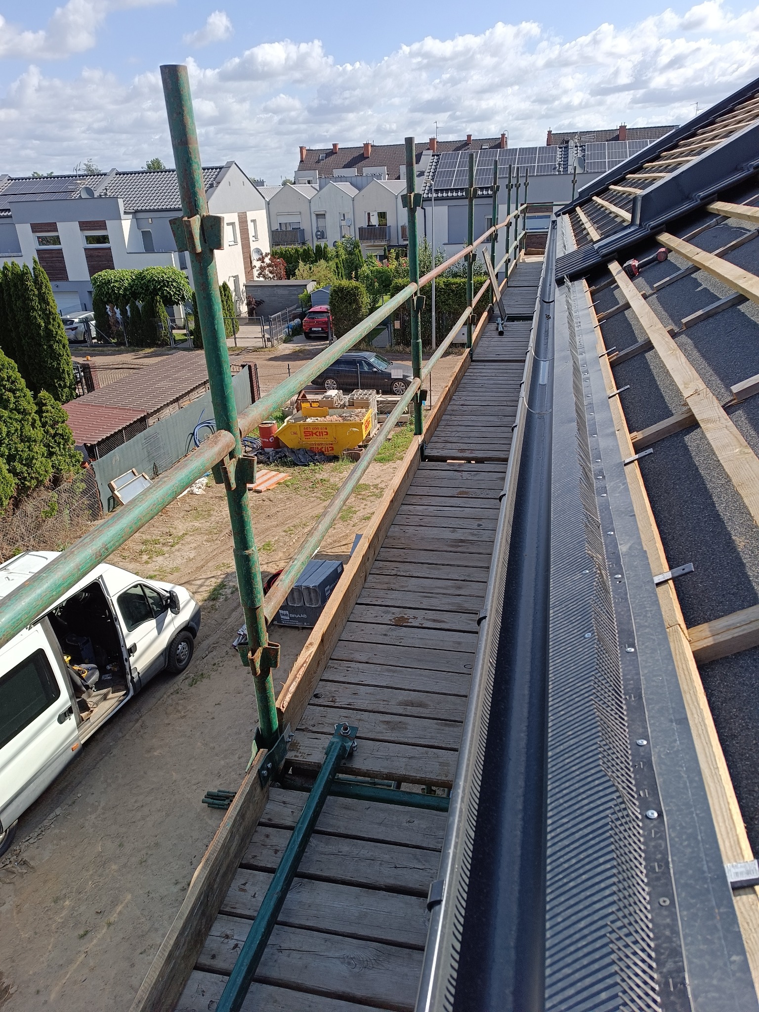 Rynna z zabezpieczeniem na dachu z rusztowaniem, widok z góry. W tle zabudowa jednorodzinna i błękitne niebo.