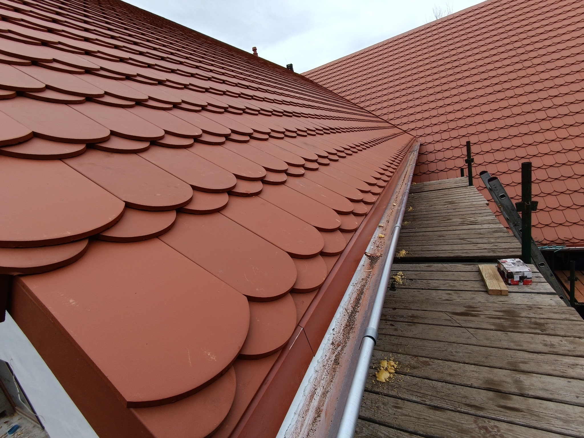 Nowy, brązowy dach z dachówki karpiówki, widok z rusztowania z rynną. Elementy montażowe i drabina w tle. Ujęcie z góry, pochmurne niebo.
