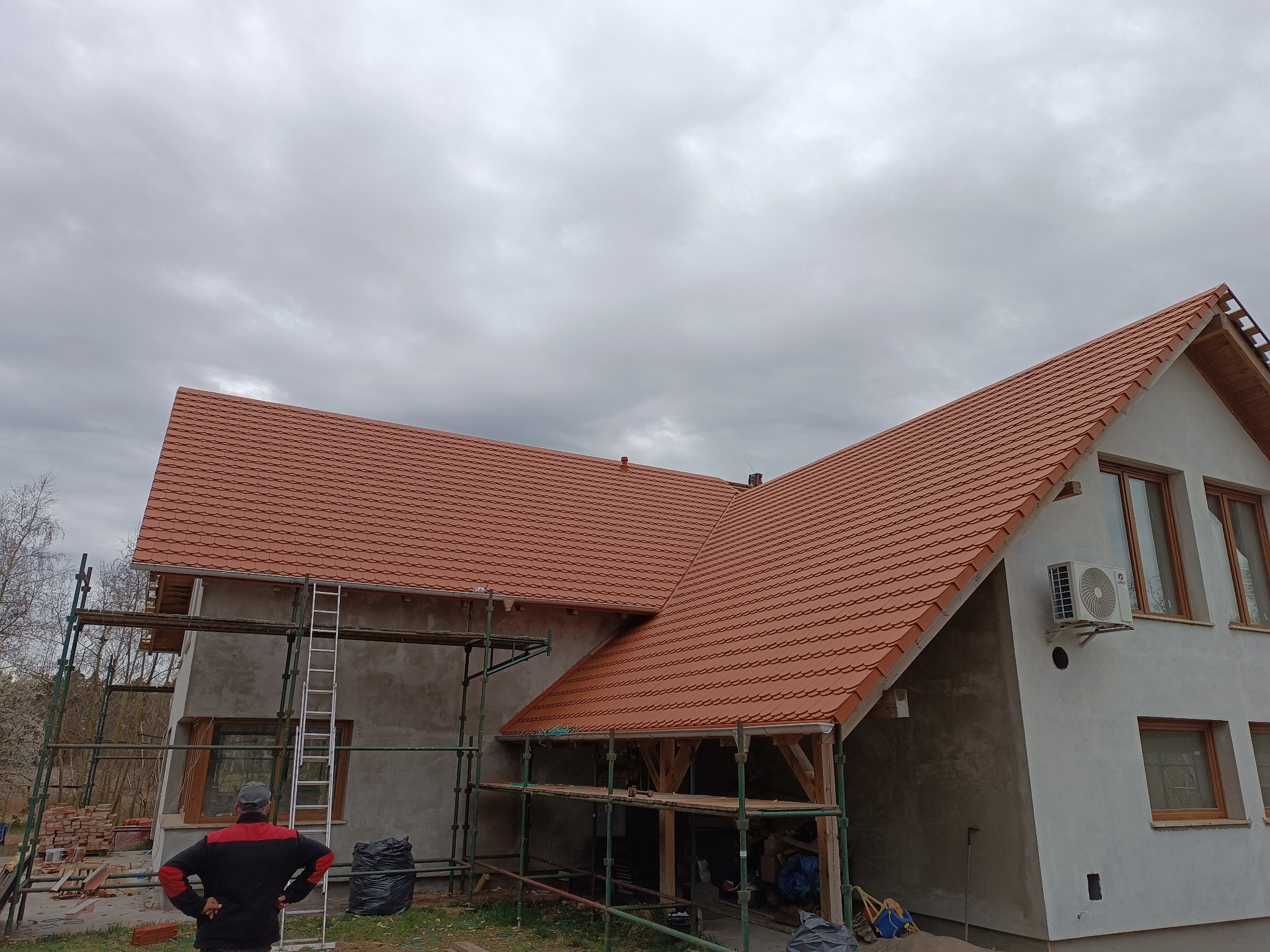 Dom w trakcie budowy z nowym, czerwonym dachem z dachówki. Widoczne rusztowania, drabina i pracownik z tyłu. Pochmurne niebo w tle.