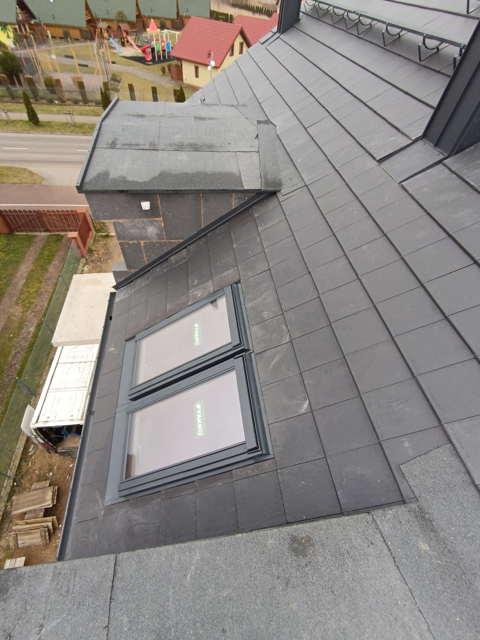 Ciemnoszary dach z dachówki ceramicznej z dwoma oknami dachowymi marki Fakro. Widok z góry na ukończony dach i okolicę z zabudową jednorodzinną.