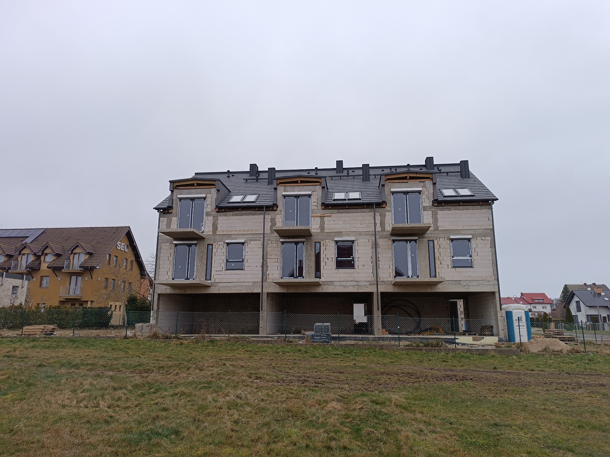 Surowy stan budynku wielorodzinnego z szarą dachówką i oknami, widoczny z zewnątrz na tle pochmurnego nieba i terenu budowy.