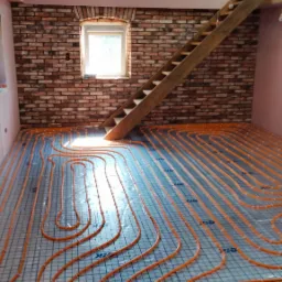 Pomarańczowe rury ogrzewania podłogowego ułożone na siatce w pomieszczeniu z ceglaną ścianą i drewnianymi elementami konstrukcyjnymi, widoczne schody.