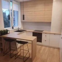 Jasna kuchnia z drewnianymi frontami szafek górnych i białymi dolnymi, blat w kształcie litery U z barkiem i czarnymi hokerami, minimalistyczne czarne lampy wiszące, drewniana podłoga.