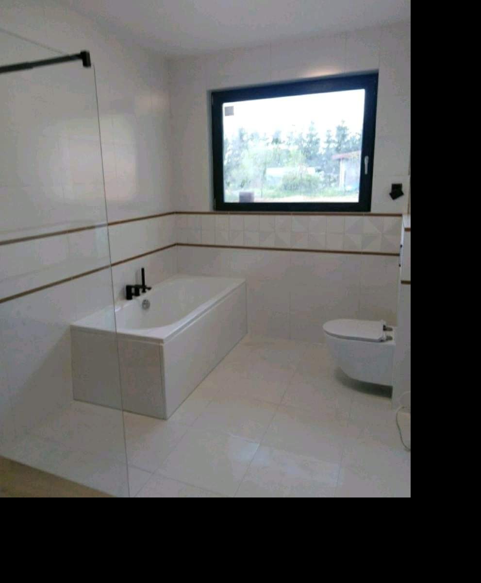 Nowoczesna łazienka w bieli z czarnymi akcentami: prostokątna wanna z czarną armaturą, podwieszana toaleta, białe płytki z dekoracyjnym paskiem, czarna rama okienna z widokiem na zieleń.