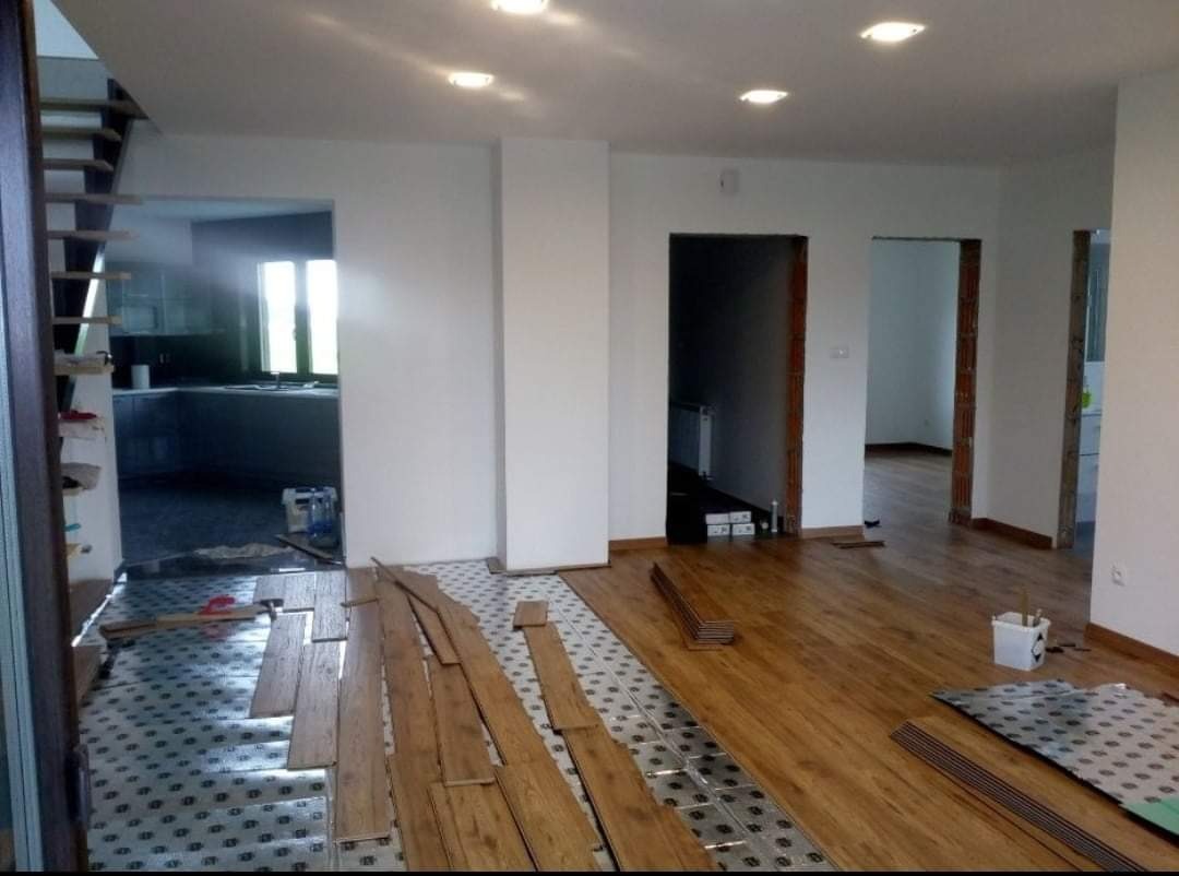 Wnętrze w trakcie prac wykończeniowych: układanie drewnianej podłogi na folii izolacyjnej, widoczne przygotowane deski, białe ściany i otwarte przejścia do innych pomieszczeń.