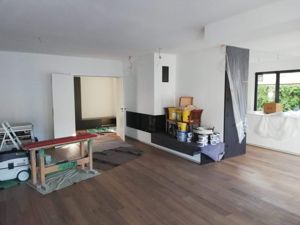 Wnętrze w trakcie remontu: widoczna podłoga z paneli, przygotowane farby i narzędzia malarskie, zabezpieczony kominek folią, drabina i odkurzacz przemysłowy w tle, częściowo pomalowane ściany...