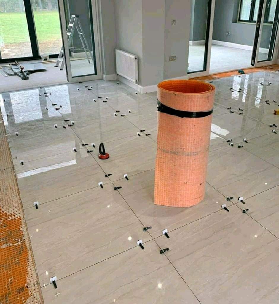 Podłoga z jasnych, błyszczących płytek, w trakcie układania z użyciem krzyżyków dystansowych i przyssawki do płytek, widoczna rolka membrany uszczelniającej w pomarańczowym kolorze.