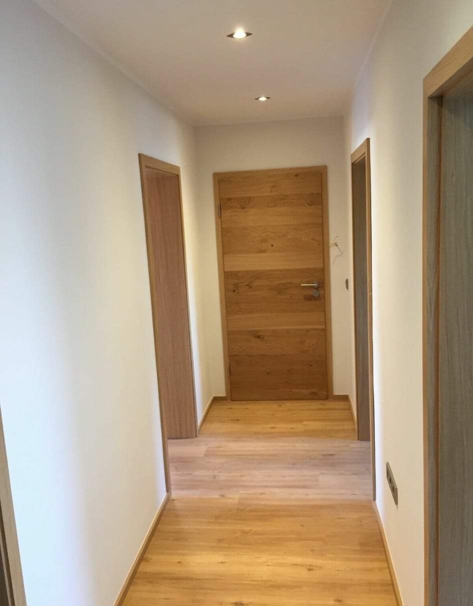 Wnętrze korytarza z trzema drewnianymi drzwiami o różnym wykończeniu, jasną podłogą z paneli i punktowym oświetleniem sufitowym.