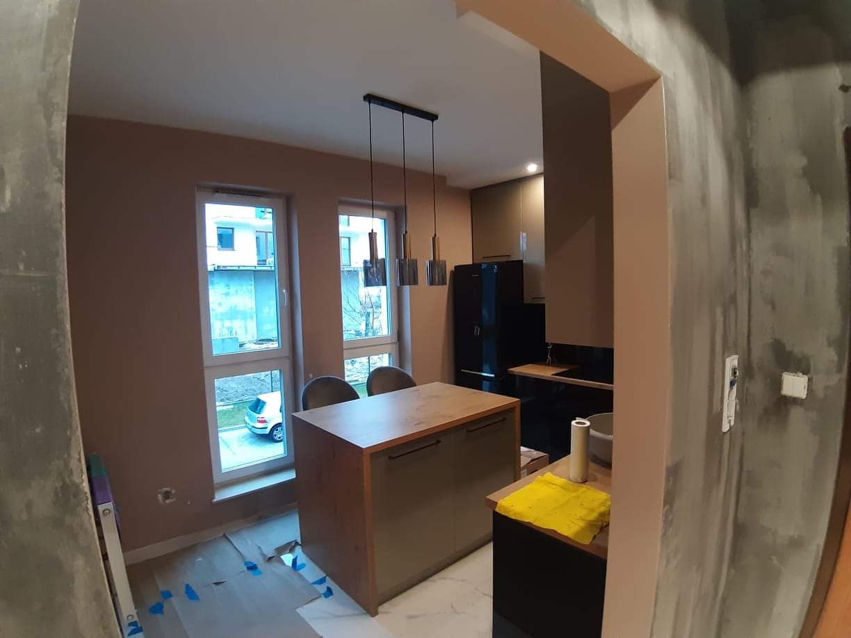 Wnętrze w trakcie prac wykończeniowych: widok na kuchnię z wyspą, ściany częściowo pomalowane na beżowo, surowy beton na ścianie obok, zabezpieczona podłoga, okna z widokiem na budynek...