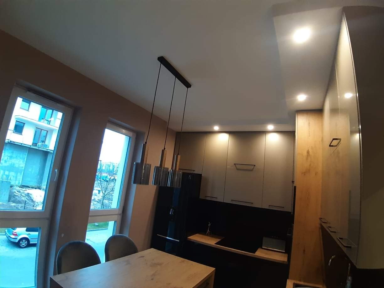 Nowoczesna kuchnia z szarymi szafkami, czarnym panelem nad blatem, drewnianym blatem i wiszącymi lampami z metalowymi elementami, widok z niskiego kąta.