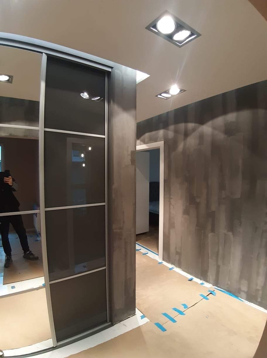 Korytarz w trakcie prac wykończeniowych: ściany pokryte szarą farbą o nieregularnej fakturze, zabezpieczona podłoga z widocznymi niebieskimi taśmami, przesuwne drzwi do szafy wnękowej z ciemnymi...