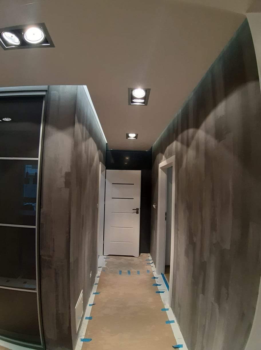 Korytarz w trakcie szpachlowania ścian na szaro, z zabezpieczoną podłogą kartonem i niebieską taśmą, widoczne drzwi i oświetlenie sufitowe.