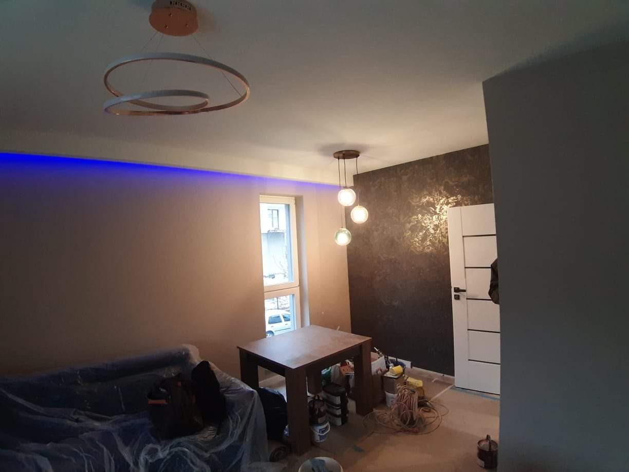 Pokój po remoncie z dekoracyjną ścianą w odcieniach brązu i złota, nowoczesnym oświetleniem sufitowym, niebieskim oświetleniem LED przy suficie i częściowo zasłoniętym meblami pod folią ochronną.