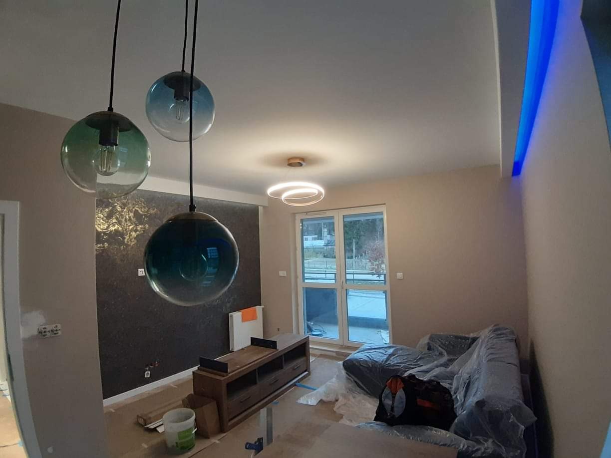 Wnętrze salonu w trakcie prac wykończeniowych: widoczne wiszące lampy kuliste w odcieniach zieleni i błękitu, dekoracyjna ściana z wzorem, meble zabezpieczone folią, podłoga częściowo zakryta...