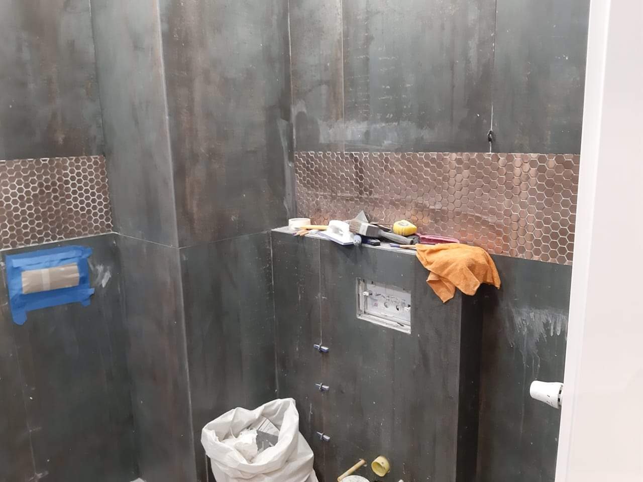 Łazienka w trakcie wykańczania z ciemnymi płytkami i mozaiką, widoczne narzędzia i materiały budowlane, zabezpieczony fragment ściany niebieską taśmą malarską.