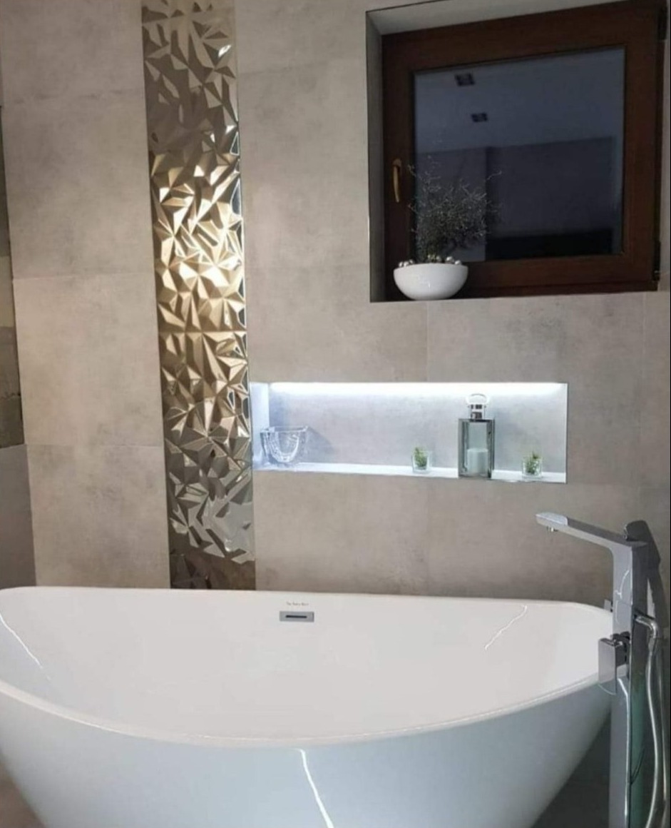 Nowoczesna łazienka z wolnostojącą białą wanną, dekoracyjną ścianą z trójwymiarowych płytek w odcieniach złota i szarości, oraz wnęką z podświetleniem LED i ozdobnymi elementami.