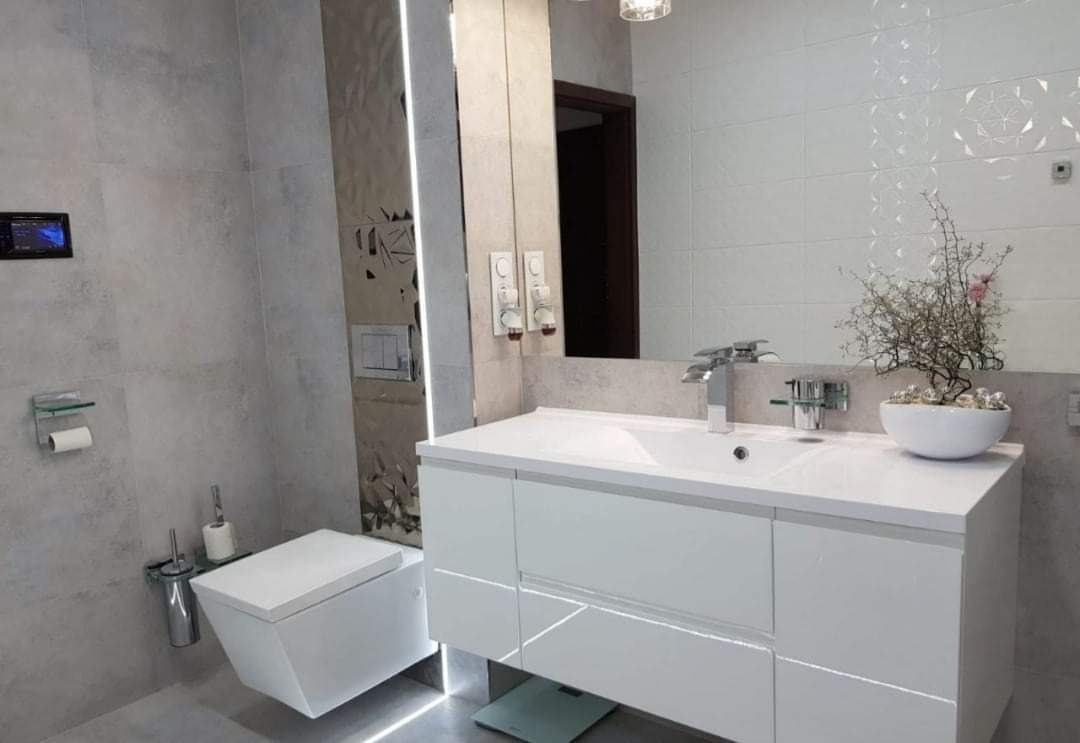 Nowoczesna łazienka z podświetlaną szafką pod umywalką, wiszącą toaletą i dekoracyjną ścianą z lustrem.