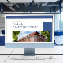 Monitor iMac wyświetlający stronę internetową osiedla Klonowe Kędzierzyn-Koźle z hasłem 'Żyj chwilą w swoim apartamencie', widok z perspektywy biurka w nowoczesnym biurze z błękitnymi akcentami.