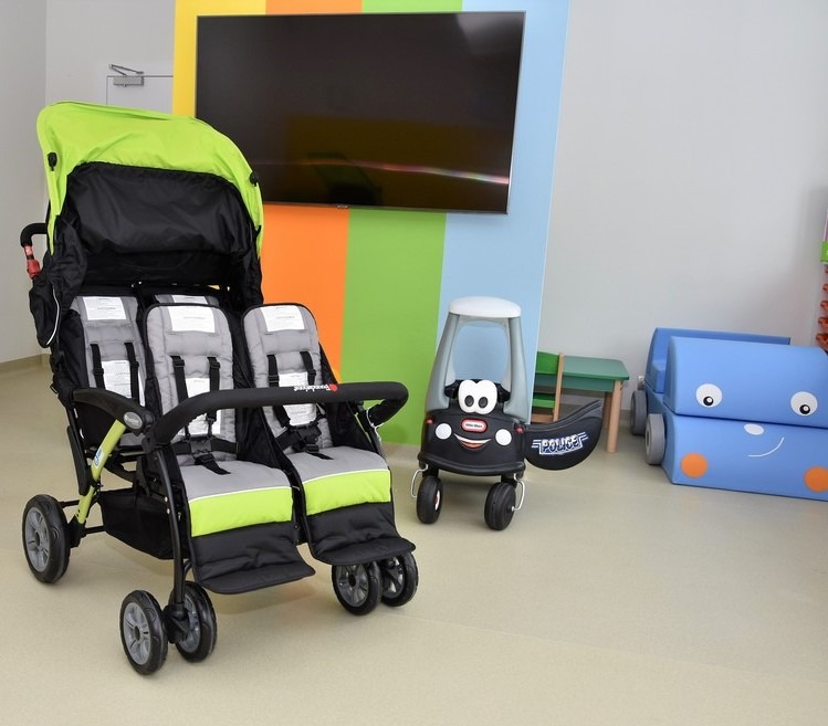Wielofunkcyjny wózek bliźniaczy w kolorach szaro-zielonym, zabawkowy samochodzik policyjny z uśmiechniętą buzią i niebieski puf w kształcie auta na tle ściany z kolorowymi pasami i telewizorem...