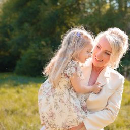 PFH KATARZYNA SOŁTYSIAK - Uśmiechnięta blond mama w kremowym żakiecie trzyma na rękach córeczkę w kwiecistej sukience na tle zieleni. Delikatne światło podkreśla radosny nastrój.