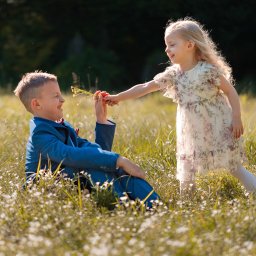 PFH KATARZYNA SOŁTYSIAK - Chłopiec w niebieskim garniturze siedzi na łące, przyjmując bukiecik od dziewczynki w kwiecistej sukience. Słoneczny dzień, naturalne światło, komunia.