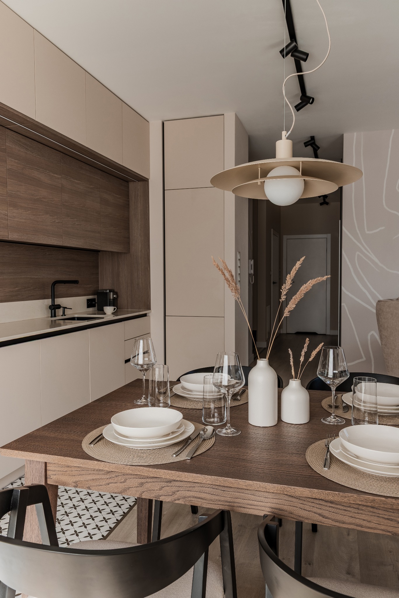 Jadalnia w neutralnych barwach: stół z ciemnego drewna, biała zastawa, szklane naczynia, minimalistyczne wazony z suszem i designerska lampa wisząca.